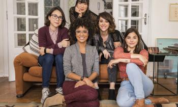 Foto do elenco de As Five. São cinco jovens mulheres: uma é negra, usa óculos e tem cabelos cacheados na altura do pescoço, três são brancas de cabelos lisos e castanhos na altura dos ombros. Uma delas usa óculos. A última mulher tem olhos puxados, pele clara, franja e cabelos lisos pretos e compridos. Elas vestem roupas casuais, cada uma com um estilo diferente, e estão sentadas em uma sala sorrindo para a câmera.