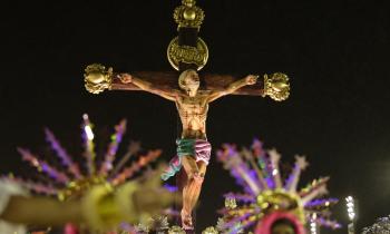  Foto de uma das alegorias apresentadas pela Estação Primeira de Mangueira durante o desfile das escolas de samba do Rio de Janeiro, no Carnaval de 2020. O carro alegórico faz uma releitura da história de Jesus Cristo, como um jovem negro baleado. Na imagem, o jovem está crucificado e olha para o céu. Acima, na cruz, lê-se a palavra "negro". Abaixo, aparecem membros da escola fantasiados, em primeiro plano mas desfocados. 