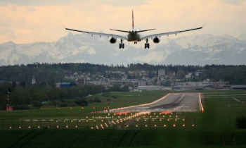 Foto de avião pousando em uma pista de aeroporto. Observa-se a parte traseira do avião, bo centro da imagem, que é branco e possui detalhes em laranja. A pista de pouso está imulinada com luzes amarelas e laranjas e ao redor há terrenod com grama e árvores baixas. Em segundo plano, há uma uma cidade, onde nota-se prédios e árvores e, ao fundo, diversas montanhas.