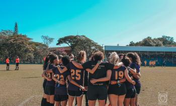 Foto de equipe esportiva reunida no Juca 2022. Na imagem, um grupo de mulheres está reunido em círculo, abraçando umas às outras. As jovens usam uniforme esportivo roxo com detalhes amarelos e shorts pretos. Elas estão em um campo com gramado. Ao fundo estão os juízes da partida e a equipe adversária. O céu azul, sem nuvens, também é evidente na imagem.