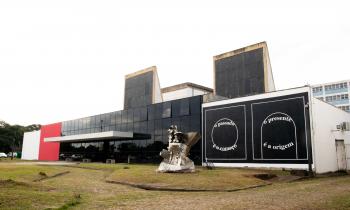 Foto da fachada do Espaço das Artes. Prédio térreo e retangular. à esquerda, uma pintura de formas geométricas, dois quadrados e circulos na cor branca em fundo preto, ao centro, o prédio possui vidro preto e na ponta, uma parede na cor vermelha. Uma obra de arte em metal está a frente do prédio.