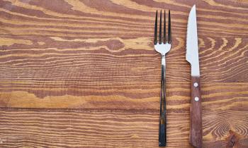 Foto de um garfo e uma faca paralelos, na vertical, alinhados à direita sobre uma superfície de madeira