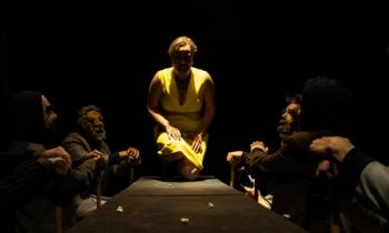 Foto de cena do espetáculo oresteia.br. Uma mulher está semi ajoelhada sobre uma mesa cercada por homens vestindo terno e usando máscaras. A mulher usa um vestido amarelo, tem pele clara e cabelos cacheados e curtos. A luz principal incide do teto, criando sombras e contrastes intensos.