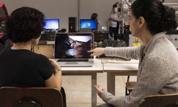 Uma mulher sentada de costas e outra sentada de lado estão lado a lado e olhando para a tela de um notebook que está sobre uma mesa. A mulher à direita aponta com o dedo indicador para a imagem que está na tela. ao fundo, há outros computadores e pessoas conversando