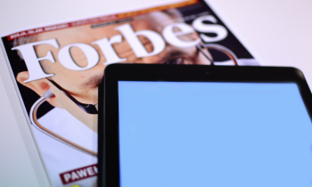 Foto de uma revista e um tablet sobre uma mesa. Em primeiro plano, o tablet, com a tela em branco. Sob o tablet, uma edição da revista Forbes. 