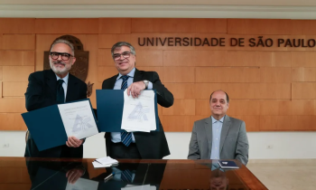 Foto do professor Paulo Nassar, um homem branco de cabelos e barba branca e óculos, e do reitor Carlos Gilberto Carlotti Jr, um homem branco de óculos e cabelos grisalhos, posando com exemplares do convênio entre ECA e Aberje. Ambos estão de pé atrás de uma mesa de madeira. Sentado na ponta da mesa, o professor Eugênio Bucci, um homem branco e calvo de cabelos escuros. Todos usam paletó e sorriem para a câmera.