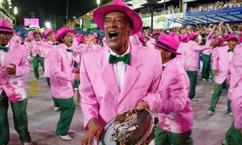 Foto de uma ala de sambistas de carnaval, com destaque para um homem negro, mais velho, tocando pandeiro e  cantando. Ele usa um terno rosa, gravata borboleta verde, camisa branca e chapéu rosa. O pandeiro tem uma imagem estampada. Os outros sambistas em volta cantam e usam a mesma roupa, incluindo uma calça verde e sapatos verde e rosa.
