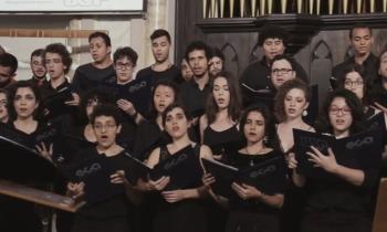 Foto de parte do Coral da ECA-USP, onde estão agrupadas várias fileiras de homens e mulheres cantando com uma pasta nas mãos