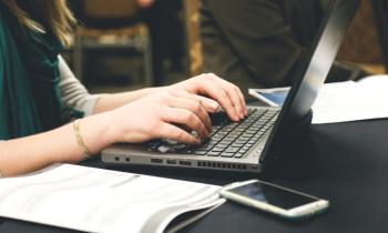 Foto das mãos de mulher branca sentada enquanto digita em um notebook cinza que está em cima de uma superfície escura. Parte de seu cabelo claro e roupa aparecem na imagem. Ao lado do notebook há um celular e um documento. 