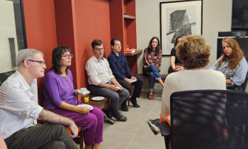 Foto de pessoas reunidas em um semi-circulo, parte delas sentada em um banco de madeira e encostados em um parede vermelha, outra parte em cadeiras de escritório. São três homens e quatro mulheres.