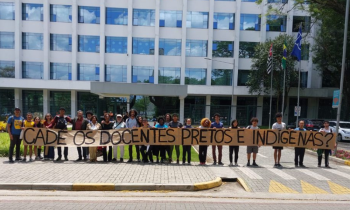  Foto de estudantes e professores em frente ao prédio da reitoria da USP. Vários estudantes, brancos e negros, estão alinhados lado a lado e seguram um cartaz bege em que está escrito "Cadê os docentes pretos e indígenas?”. Atrás dos estudantes, há um prédio espelhado