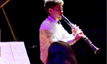 Foto de um homem tocando clarinete. Ele é branco, tem cabelos curtos e escuros e veste uma camisa branca. O fundo da imagem é totalmente preto, e o jovem é iluminado por luzes branca e roxa. Na porção inferior da foto, aparecem a cabeça de uma mulher que está de costas para a câmera e uma partitura apoiada no piano que ela toca.