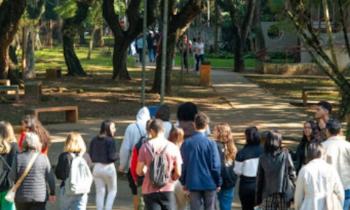  Foto tirada durante a visita monitorada à ECA. Na imagem, um grupo de estudantes passa pela parte externa da unidade, onde há várias árvores e bancos de concreto. Na foto, conta-se 19 pessoas andando de costas para a câmera.