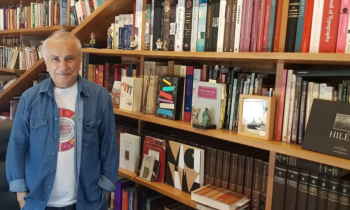 Foto de um homem posicionado em frente a uma estante de livros. Ele é branco, tem cabelos curtos e grisalhos e olhos escuros. Ele veste uma camiseta branca estampada e uma camisa jeans por cima. A estante é grande e ocupa toda a parede e, consequentemente, todo o fundo da imagem.