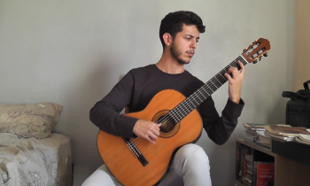 Foto de um rapaz tocando violão clássico e olhando para o instrumento. Ele é branco, tem cabelos curtos e escuros e veste uma blusa de manga longa preta e calça cinza. O jovem está em um quarto, sentado entre uma cama e uma escrivaninha. A parede ao fundo é cinza.