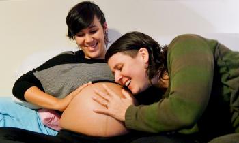 Foto de duas mulheres sentadas. A da esquerda é branca, tem cabelos curtos e escuros e veste uma blusa cinza e preta. Ela está grávida e sua barriga está exposta. A mulher da direita é branca, tem cabelos curtos e escuros, veste uma blusa verde e com o rosto próximo à barriga da companheira. As duas estão sorrindo e com uma das mãos na barriga grávida.