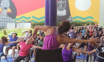 Foto de atividade no Sesc Pinheiros. Grupo de mulheres praticam atividade física. A professora está sentada em uma cadeira, de costas. Ela tem cabelos castanhos, ligeiramente ondulados, presos no topo da cabeça. Ela veste uma blusa roxa regata. à frente dela, está um grupo de mulheres sentadas em cadeiras, uma ao lado da outra. Elas repetem os movimentos da professora: um braço se estica para o lado, torcendo o tronco para o mesmo lado. o outro braço se mantém dobrado. Ao fundo, um painel artístico.