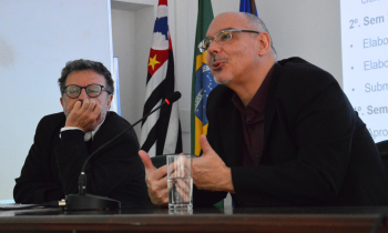 Foto de dois homens sentados em uma mesa. À esquerda, um homem de cabelos levemente grisalhos, usando óculos com haste escura, um terno preto e uma camisa branca, está com o queixo apoiado em uma das mãos. À direita, um homem calvo, de barba rala, usando óculos, um paletó preto e uma camisa escura fala ao microfone e gesticula com as mãos. Ao fundo, uma parede branca e as bandeiras do estado de São Paulo, do Brasil e da USP.