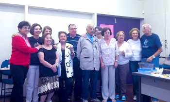 Foto de nove mulheres e três homens reunidos posando para uma foto de grupo. São todas pessoas idosas. Atrás das pessoas há uma porta dupla fechada, uma parede e é possível ver uma carteira de sala de aula. Também é possível ver uma mesa de professor com algumas vasilhas e sobras de comida. Aparentemente se trata de uma confraternização.