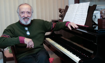 Foto de um homem branco, calvo, de cabelos e barba brancos. Ele usa um suéter verde escuro com punhos vermelhos e calça preta. Seus óculos de grau estão apoiados na gola do suéter. Ele sorri e posa ao lado de um piano de madeira escura, sobre o qual estende o braço esquerdo. Sobre a tampa do piano há uma partitura aberta..Ao fundo, uma persiana bege.