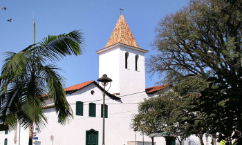 Foto da fachada de uma igreja em estilo colonial. As paredes são brancas, enquanto portas e janelas são da cor azul-marinho. Ao centro, a torre, que possui duas janelas estreitas e um telhado em forma de pirâmide com uma cruz no topo. A base da torre se liga, pelas laterais, a duas casas. Em primeiro plano, um poste de luz e as folhagens de uma árvore e de uma palmeira. Ao fundo, o céu azul, sem nuvens. 