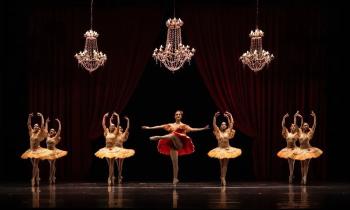 Foto de um espetáculo de ballet. Na imagem, oito bailarinas vestem roupas customizadas. No lado esquerdo, com quatro bailarinas, e no lado direito, com três bailarinas, elas vestem figurinos amarelos. A bailarina no centro usa um figurino vermelho. Atrás delas há um fundo escuro e coxias vermelhas, acima existem três lustres. 