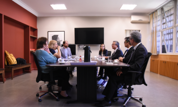 Foto de uma mesa de reuniões, com pessoas sentadas em cadeiras em volta dela. Ao fundo, um monitor de tv grande. Sobre a mesa, copos, xicaras e uma garrafa de café.