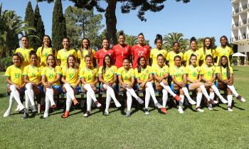 Foto oficial da Seleção Brasileira Feminina de 2019. 25 mulheres posam para uma foto, sendo 12 em pé e, à frente delas, 13 sentadas em um banco com as pernas cruzadas. Três das mulheres, que estão em pé e no centro da fotografia, usam um uniformes vermelhos, as demais usam uniformes amarelo e azul e meião branco. Elas estão em um gramado, a maioria sorri para a câmera e há um prédio branco de quatro andares no canto direito da foto. Árvores de diferentes formatos formam o segundo plano da imagem. 