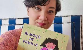 Foto da escritora Janaína Tokitaka, uma mulher de traços asiáticos, olhos castanhos, cabelos grisalhos, curtos e ondulados. Ela olha em direção à câmera e, com uma das mãos, segura seu livro Almoço de Família, cuja capa tem a ilustração de uma menina com cabelos cacheados e escuros, vestindo  blusa cor de rosa, em frente a uma mesa com pratos de comida. Ao lado da menina, na ilustração, há um gato.  Atrás de Janaína há um estofado com listras azuis e brancas e uma parede branca.