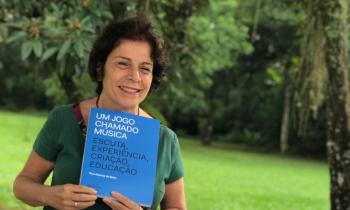 Foto de uma mulher branca de cabelos curtos e escuros. Ela segura um livro com capa azul de título Um Jogo Chamado Música: escuta, experiência, criação, educação enquanto sorri e olha para o lado direito da imagem. Ao fundo, um gramado e folhagens de árvores. 