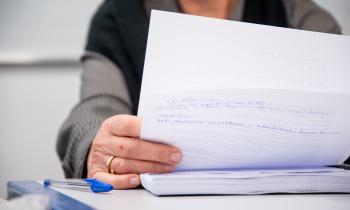 Foto de uma professora durante uma banca avaliadora. A foto destaca a mão da professora, segurando duas folhas de papel, no verso da folha, algumas frases escritas a caneta. Ela apoia o braço na mesa e sobre uma tese, um caderno espiral grosso. Na mão, ela usa uma aliança. Ao lado, sobre a mesa, uma caneta azul. Mais ao fundo, podemos observar o braço da professora, levemente desfocado. Ela veste uma blusa de lã nas cores cinza e preto.