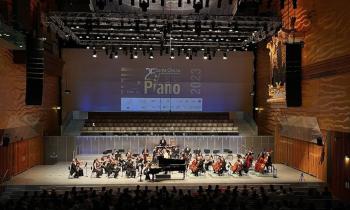 Foto do palco do 25o Concurso Internacional de Piano Santa Cecília. Na imagem, uma orquestra se apresenta para um público em uma sala de concerto. Ao centro do palco, um piano grande e preto. Ao fundo, no telão, aparece o cartaz de divulgação do concurso. A iluminação é direcionada ao palco e as paredes do ambiente têm tons de marrom e laranja. 