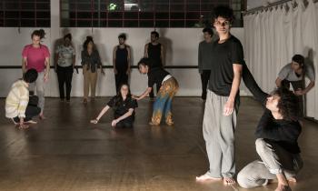 Foto. Em uma sala ampla com piso de madeira, paredes brancas, vitrô ao fundo e uma cortina branca no canto direito, um grupo de pessoas participa de uma aula de dança e expressão corporal. À frente, estão dois homens, jovens, brancos, com cabelos castanhos e ondulados; um está de pé e olha para baixo com expressão séria, o outro está agachado, erguendo um dos braços e tocando nas costas do primeiro. Eles se vestem de forma semelhante: calça cinza e blusa preta, descalços