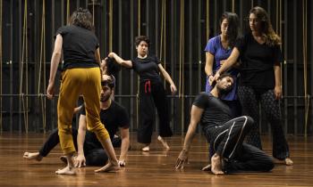 foto de sete jovens movendo-se em um palco. Eles são, na maioria, brancos,estão descalços e usam calças e camisetas de tecidos confortáveis e modelagem larga. A maioria das roupas é da cor preta, destacando-se uma calça amarela, uma camiseta azul e uma fita vermelha. Dois jovens estão semi ajoelhados, com as mãos tocando o piso, enquanto outros tocam suas cabeças com os braços esticados.  O piso é de madeira e no fundo escuro estão as cordas verticais que fazem a mecânica cênica.