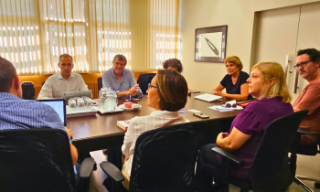 Foto de um grupo de pessoas sentado ao redor de uma mesa retangular. Na frente, de costas ou de lado para a foto, estão os docentes da ECA: dois homens e duas mulheres, sentados em cadeiras de escritório, na cor preta. Ao fundo, e de frente para a foto, estão três docentes da faculdade francesa. Na cabeceira da mesa, está sentada a diretora da ECA. Em cima da mesa de madeira, na cor marrom escuro, estão diversos objetos, jarras de água, copos de vidro, computadores, papeis, livros, jarra de café.