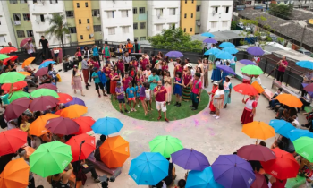 Foto de diversas pessoas reunidas na área externa de um conjunto habitacional. Elas formam uma roda, com diversas pessoas usando roupas coloridas e portando guarda-chuvas nas cores azul, roxo, verde, vermelho, vinho e laranja. No centro da roda, um jovem branco de cabelos escuros, camiseta vermelha e bermuda branca fala e gesticula. Ao lado dele há crianças e jovens, muitos deles negros. Ao fundo, parte da fachada de alguns prédios. 