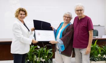 Três pessoas posam de pé para uma fotografia. Duas mulheres seguram uma pasta preta aberta, uma de cada lado, exibindo um certificado. À esquerda, está uma mulher loira, de cabelos curtos, vestindo um blazer branco, camisa e calças pretas. Do outro lado, está uma mulher branca, de cabelos brancos, lisos e curtos. Ela usa óculos de haste avermelhada, veste calça e blusa brancas e uma blusa, do tipo quimono, vermelho escuro com detalhes em azul claro. Do lado direito da imagem, abraçando a segunda mulher, est