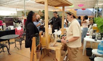 Foto de uma feira em um pátio coberto. Em primeiro plano, estão duas pessoas conversando em pé separadas por uma banca de madeira com utensílios para fazer café. A pessoa da esquerda é negra, possui cabelos médios, castanhos e cacheados. Ela usa óculos, blazer preto e camisa branca. Do outro lado da banca, há uma pessoa branca, de cabelos curtos e castanhos. Ela usa chapéu cor de palha, camisa branca e calça bege. Ao fundo, há mais bancas, com produtos e pessoas circulando, cadeiras de plástico e plantas. 