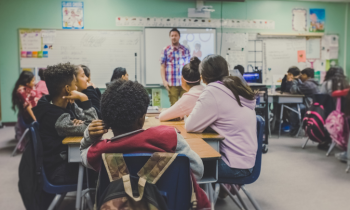 Fotografia de uma sala de aula com parede verde, piso cinza, quadro branco e cartazes. Estudantes  estão sentados em grupos em carteiras dispostas na sala com a atenção voltada ao professor, um homem branco que veste camisa xadrez e está à frente do quadro branco. 