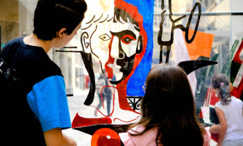 Uma criança de cabelos compridos e blusa Rosa e uma joem de cabelos curtos e blusa preta estão em pé, de costas, observando uma obra de arte, o desenho do busto de um homem, feito em materiais diversos, nas cores preto, vermelho azul e transparência. Ao fundo, prédios e a calçada mostram que elas estão em um espaço aberto da cidade de São Paulo