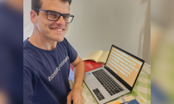 Foto de um jovem de pele branca, cabelos castanhos escuros e óculos de armação, vestindo uma camiseta azul escura. Ele está sentado e com o antebraço apoiado em uma mesa. à sua frente e em cima da mesa está um notebook e alguns livros. A parede ao fundo é branca