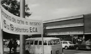 Captura de vídeo. A imagem é em preto e branco e mostra em primeiro plano, uma faixa de tecido com os dizeres Semana de Arte e Ensino. 15 a 19 de setembro. ECA. Atrás da faixa estão alguns carros e a fachada do Prédio Central da ECA, uma construção com três pavimentos marcados por três lajes horizontais intercaladas por elementos vazados à frente das janelas.