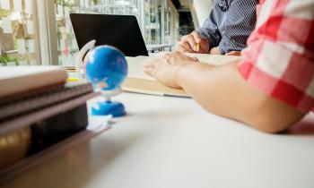 Foto de um uma mesa, sobre a qual repousam: livros e cadernos em uma pilha pequena, um globo azul, um note book e um livro. Em cima do livro, estão a mão e o antebraço de uma pessoa. Ao seu lado, outra mão segura um lapis e aponta para uma das páginas do livro aberto. No fundo, há janelas.