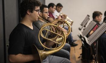 Foto de uma fileira de naipe de metais de uma orquestra, com quatro pessoas de pele clara e cabelos curtos e escuros lado a lado em frente de estantes de partituras. A pessoa em primeiro plano toca uma trompa, instrumento de sopro metálico circular, dourado. As pessoas tocando no fundo da fileira estão mais desfocadas.