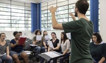 Foto de um jovem branco, de cabelos curtos e castanhos e barba da mesma cor, está de pé, com os braços ao alto. à sua frente uma partitura e mais ao fundo, um grupo de jovens estão sentados, com folhas nas mãos, cantando. Ao fundo, as janelas da sala de aula.