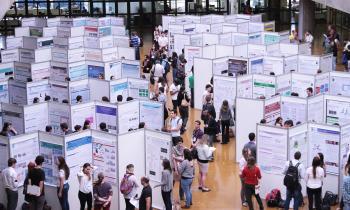 Fotografia de um espaço amplo com vários painéis expositivos do tipo biombo, organizados em duas colunas e oito filas. Diversas pessoas circulam entre os biombos, conversam entre si e observam os banners dispostos nos painéis.