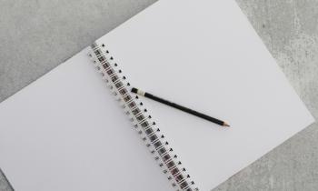 Foto de um caderno com espiral aberto e sem pauta, sobre o qual está apoiado um lápis preto com borracha em uma das pontas. O caderno está sobre um fundo cinza.