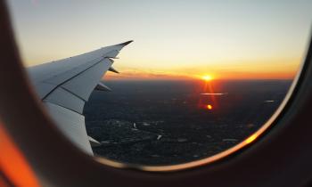 Foto da asa de um avião tirada pelo lado de dentro do avião. Na foto aparece uma extensa faixa de solo e um céu com tons de laranja e azul com o Sol no horizonte