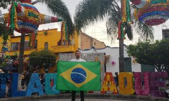 Foto de Wellington Nardes em frente a um letreiro, em letras coloridas, da cidade mexicana de Tlaquepaque. Wellington está no centro da foto, de pé, segurando uma bandeira do Brasil. É um homem branco, com cabelos castanhos, barba castanha levemente grisalha, veste camiseta branca, calça preta e tênis branco. Na parte de cima da foto, há piñatas coloridas penduradas por fios brancos. Ao fundo, há casas antigas, uma amarela e uma branca, e palmeiras. 