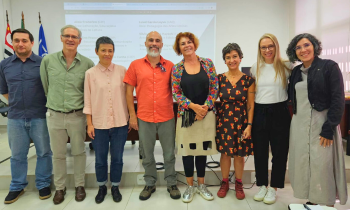 Foto de sete pessoas de pé em uma auditório, lado a lado, posando. Ao fundo, uma parede branca, com uma projeção, e as bandeiras de São Paulo, Do brasil e da USP, ao fundo, do lado esquerdo.